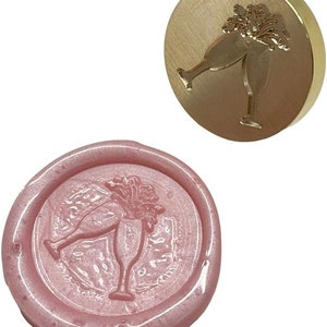 May include: A gold-toned metal wax seal stamp with a design of two champagne glasses clinking together. The stamp is used to create a wax seal with the same design, shown in pink.