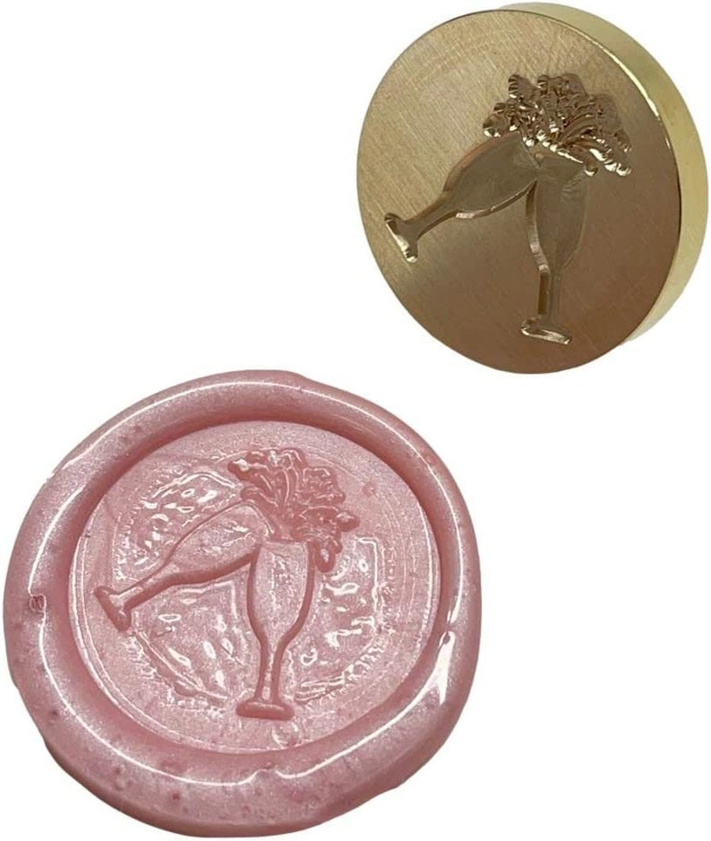 May include: A gold-toned metal wax seal stamp with a design of two champagne glasses clinking together. The stamp is used to create a wax seal with the same design, shown in pink.