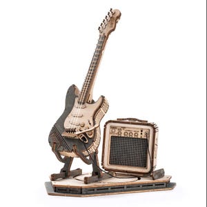 May include: A laser-cut wooden model kit of an electric guitar and a small amplifier. The guitar features a dark grey body with light wood accents, a tremolo arm, and a stand. The amplifier has a black mesh grille, control knobs, and a power switch.