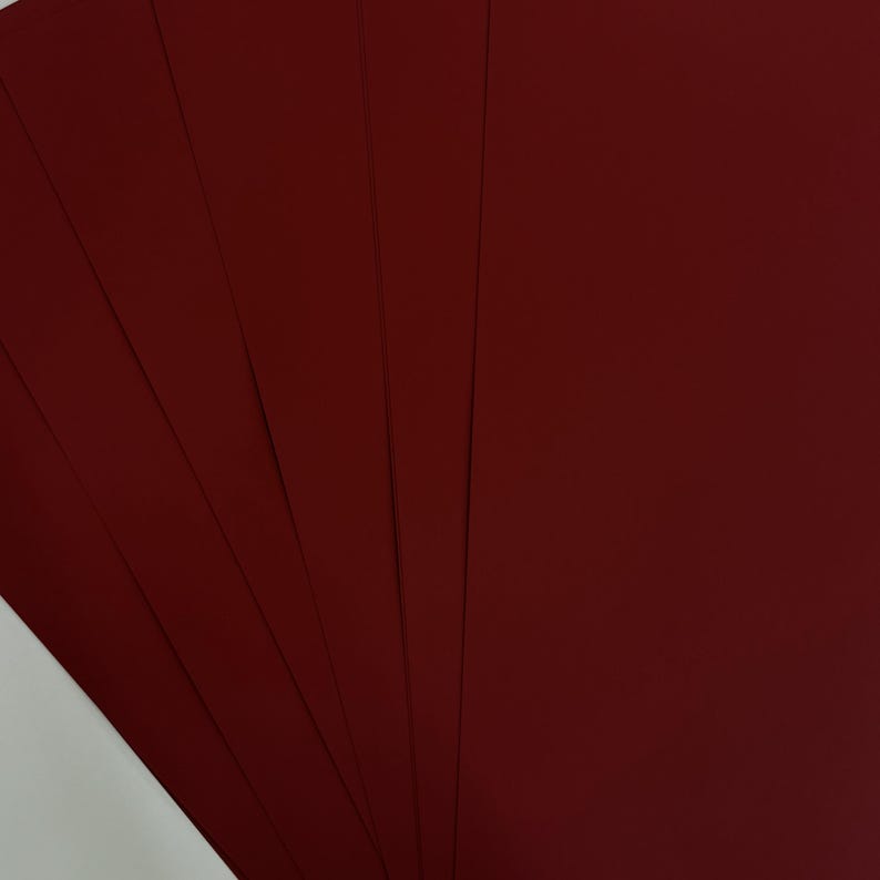 May include: A stack of burgundy-colored cardstock paper fanned out, showcasing the rich, deep red hue. The paper has a smooth, matte finish, and the edges are neatly aligned. The image highlights the color and texture of the paper.