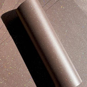 May include: A roll of brown glitter paper with a shimmering, iridescent effect. The paper is partially unrolled, revealing a stack of matching sheets. The glitter catches the light, creating a sparkling appearance, ideal for crafts and decorations.