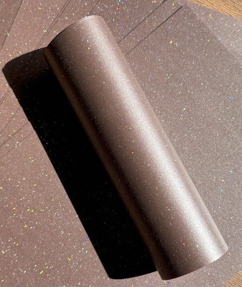 May include: A roll of brown glitter paper with a shimmering, iridescent effect. The paper is partially unrolled, revealing a stack of matching sheets. The glitter catches the light, creating a sparkling appearance, ideal for crafts and decorations.