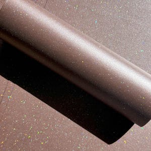 May include: A roll of brown glitter paper with a shimmering, iridescent effect. The paper is partially unrolled, revealing a flat surface covered in the same glitter. The glitter catches the light, creating a sparkling appearance.