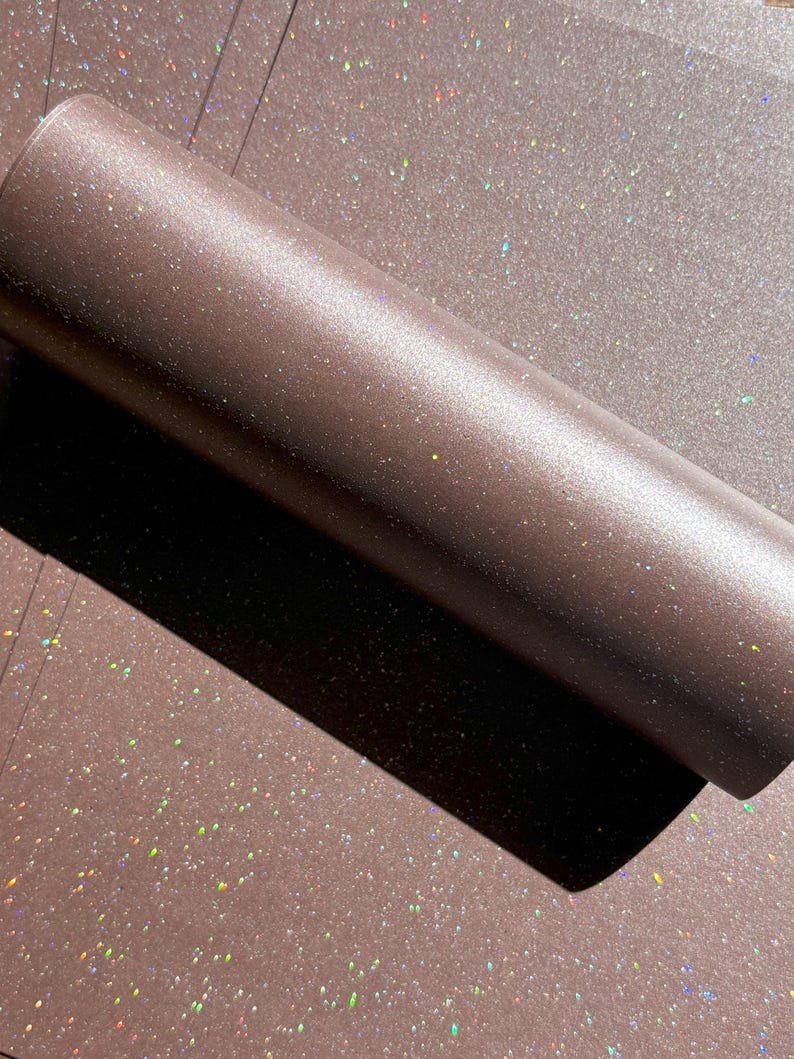 May include: A roll of brown glitter paper with a shimmering, iridescent effect. The paper is partially unrolled, revealing a flat surface covered in the same glitter. The glitter catches the light, creating a sparkling appearance.