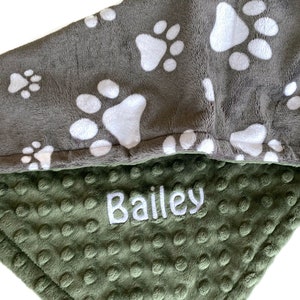 Op de afbeelding: Een grijs en groen fleece deken met witte poot afdrukken aan de ene kant en een groene minky stof aan de andere kant. De deken heeft de naam "Bailey" geborduurd op de groene kant.