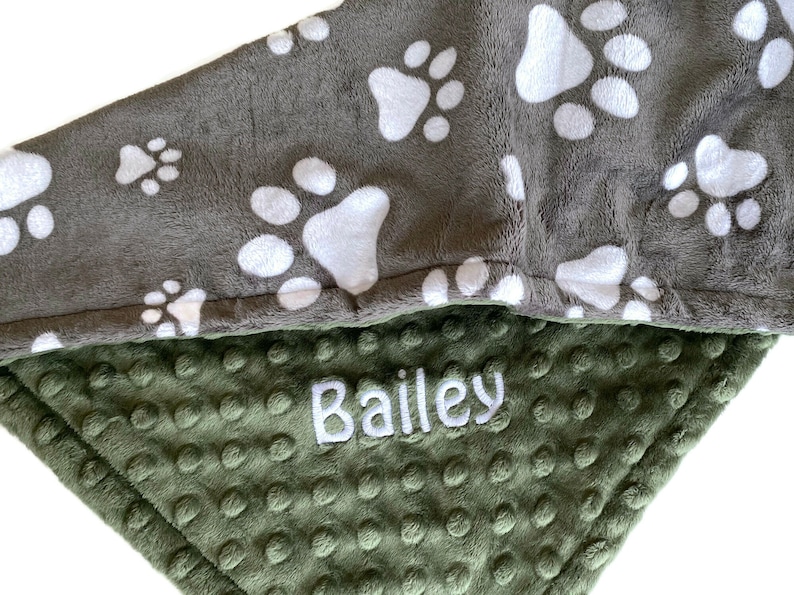 Op de afbeelding: Een grijs en groen fleece deken met witte poot afdrukken aan de ene kant en een groene minky stof aan de andere kant. De deken heeft de naam "Bailey" geborduurd op de groene kant.