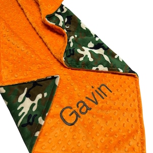 May include: An orange baby blanket with a dimpled texture and a camouflage pattern on the reverse side. The name "Gavin" is embroidered in dark thread on the blanket. The blanket is folded, showing both sides.