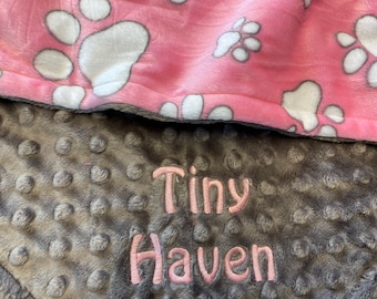 May include: A grey and pink baby blanket with a soft, textured surface. The blanket has a white paw print pattern on the pink side and the words "Tiny Haven" embroidered on the grey side.