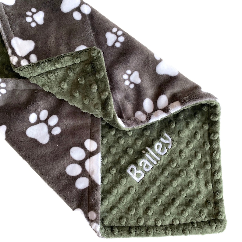 Op de afbeelding: Een groene en bruine fleece deken met een wit pootprintpatroon. De deken is gepersonaliseerd met de naam "Bailey" geborduurd op de groene kant.