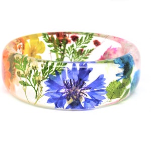 May include: A clear resin bangle bracelet with pressed flowers and leaves. The flowers are in shades of yellow, orange, pink, blue, and green.