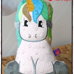 Puede incluir: Un peluche de unicornio con cuerpo rosa, patas grises y melena colorida. La melena presenta tonos de azul, verde y marr&oacute;n. El unicornio tiene ojos negros y un cuerno dorado. El texto "La Joya" es visible.