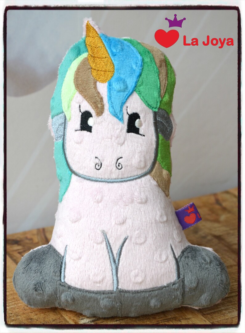 Puede incluir: Un peluche de unicornio con cuerpo rosa, patas grises y melena colorida. La melena presenta tonos de azul, verde y marr&oacute;n. El unicornio tiene ojos negros y un cuerno dorado. El texto "La Joya" es visible.
