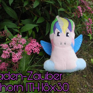 Puede incluir: Un peluche de unicornio con melena arco&iacute;ris y alas azules, sobre un fondo de flores rosas y vegetaci&oacute;n. El texto "Faden-Zauber Einhorn ITH 18x30" se muestra en fuente p&uacute;rpura.