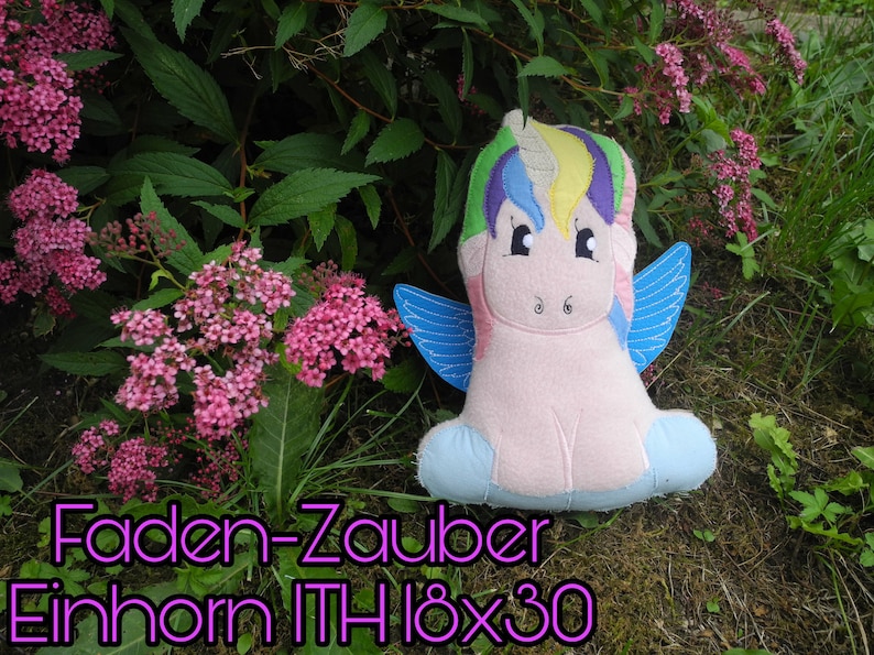 Puede incluir: Un peluche de unicornio con melena arco&iacute;ris y alas azules, sobre un fondo de flores rosas y vegetaci&oacute;n. El texto "Faden-Zauber Einhorn ITH 18x30" se muestra en fuente p&uacute;rpura.