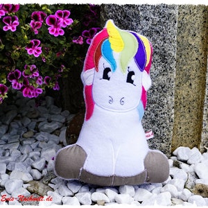 Puede incluir: Un unicornio de peluche blanco con melena y cola de arco&iacute;ris, sentado sobre piedras blancas. El unicornio tiene una cara sonriente con ojos negros y est&aacute; rodeado de flores moradas. El peluche tiene pezu&ntilde;as marrones.