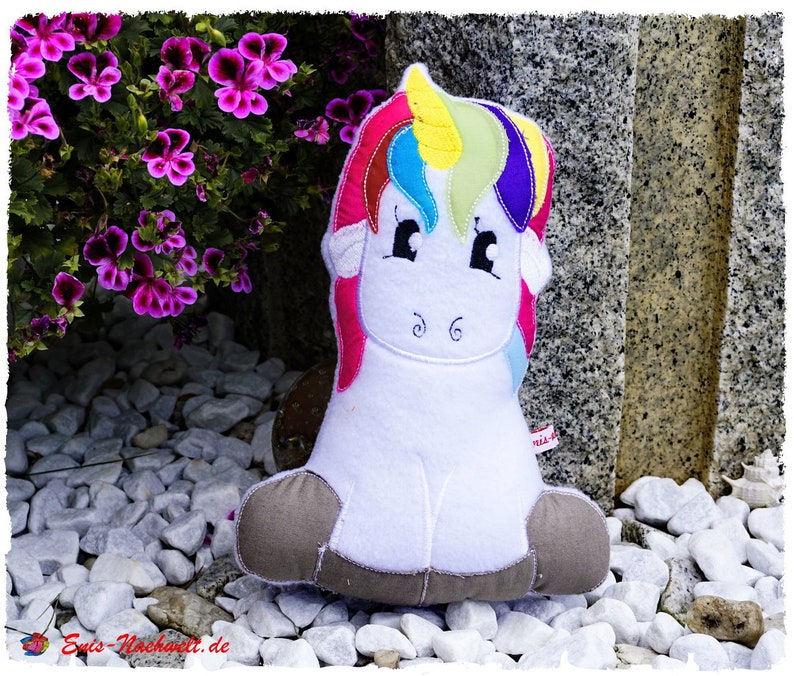 Puede incluir: Un unicornio de peluche blanco con melena y cola de arco&iacute;ris, sentado sobre piedras blancas. El unicornio tiene una cara sonriente con ojos negros y est&aacute; rodeado de flores moradas. El peluche tiene pezu&ntilde;as marrones.