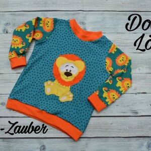 May include: A teal blue long-sleeved shirt with orange trim and a cute lion applique. The shirt has a small white polka dot pattern and features a lion with a yellow body, orange mane, and black eyes. The sleeves have a repeating pattern of small lions. The shirt has the text "Doodle L&ouml;we" and "Faden-Zauber" printed on it.