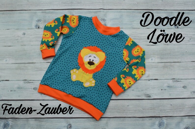 May include: A teal blue long-sleeved shirt with orange trim and a cute lion applique. The shirt has a small white polka dot pattern and features a lion with a yellow body, orange mane, and black eyes. The sleeves have a repeating pattern of small lions. The shirt has the text "Doodle L&ouml;we" and "Faden-Zauber" printed on it.