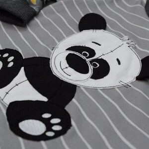 May include: A gray and white striped shirt with a black and white panda bear applique. The panda bear has a white face with black eyes and a black nose. The panda bear is wearing a white belly.