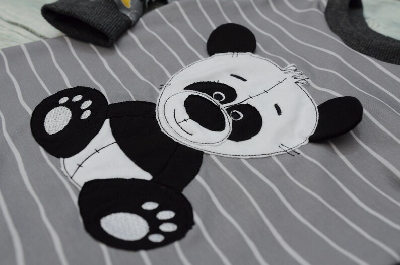 May include: A gray and white striped shirt with a black and white panda bear applique. The panda bear has a white face with black eyes and a black nose. The panda bear is wearing a white belly.