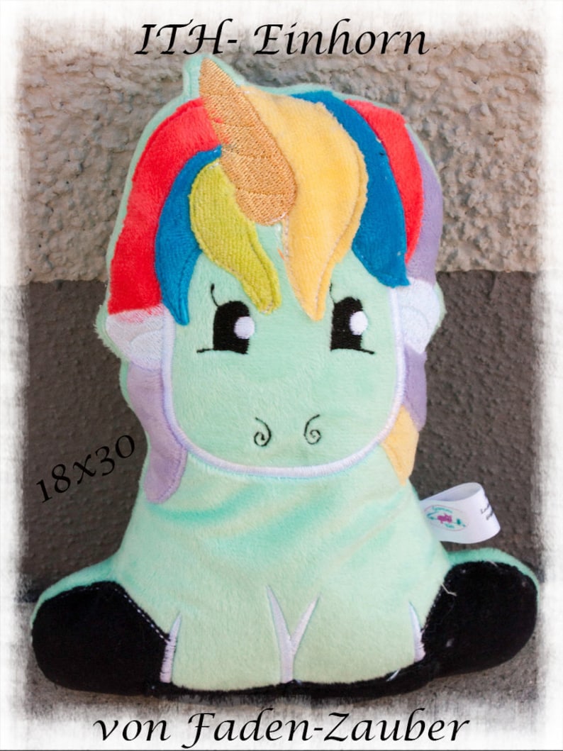 Puede incluir: Un peluche de unicornio en tonos verde menta, con una melena arco&iacute;ris y un cuerno dorado. Los textos "ITH- Einhorn" y "von Faden-Zauber" son visibles. El juguete mide 18x30 cm.