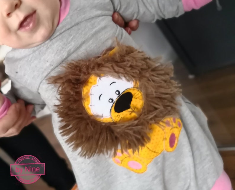 May include: A gray long-sleeved onesie with a pink trim. The onesie features an embroidered lion with a brown furry mane. The lion is wearing a yellow and orange body with pink paw prints.