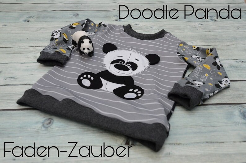 May include: A gray and black striped long-sleeved shirt with a black and white panda applique. The sleeves have a gray and white panda print with yellow accents. The shirt has the text "Doodle Panda" and "Faden-Zauber" printed on it.