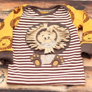 May include: A brown and white striped long-sleeved shirt with a yellow lion applique on the front. The lion has a brown face, a brown mane, and brown paws.