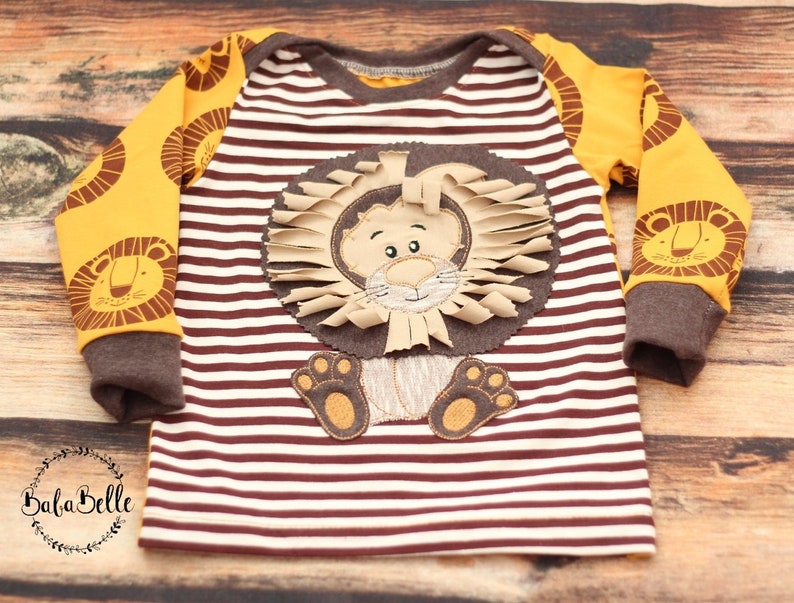 May include: A brown and white striped long-sleeved shirt with a yellow lion applique on the front. The lion has a brown face, a brown mane, and brown paws.