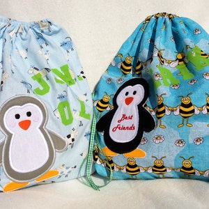 May include: Two drawstring bags. One is light blue with a penguin applique and the name "JN 01" in green. The other is turquoise with a penguin applique that says "Best Friends" and a pattern of bees.