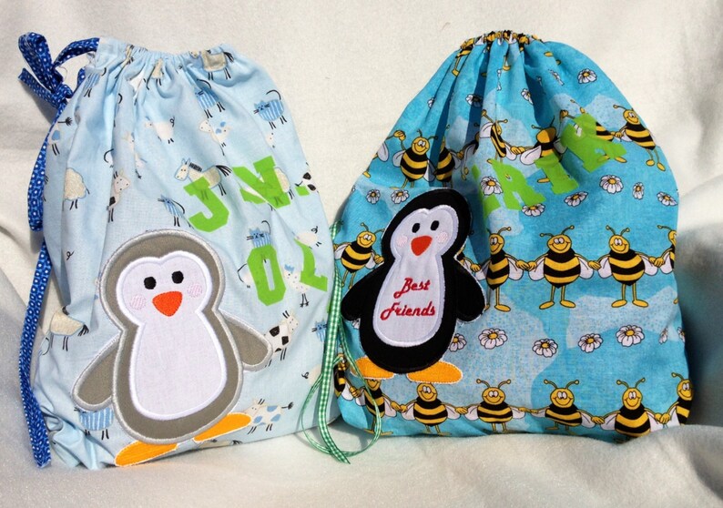May include: Two drawstring bags. One is light blue with a penguin applique and the name "JN 01" in green. The other is turquoise with a penguin applique that says "Best Friends" and a pattern of bees.