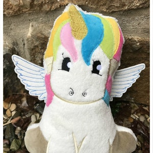 Puede incluir: Un peluche de unicornio con cuerpo blanco, melena arco&iacute;ris y cuerno dorado. El unicornio tiene ojos y alas bordadas. El texto "Einhorner ITH by Faden-Zauber" es visible en la parte superior de la imagen.