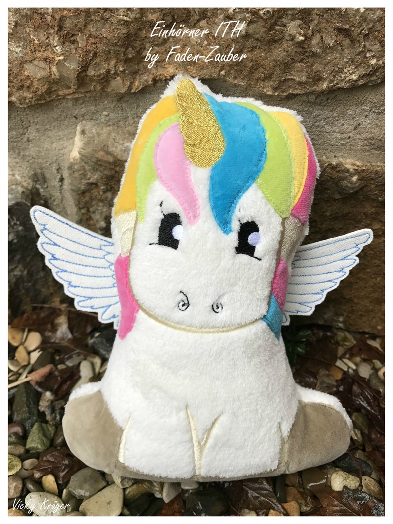 Puede incluir: Un peluche de unicornio con cuerpo blanco, melena arco&iacute;ris y cuerno dorado. El unicornio tiene ojos y alas bordadas. El texto "Einhorner ITH by Faden-Zauber" es visible en la parte superior de la imagen.
