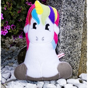 May include: A plush unicorn toy with a white body, gray hooves, and a colorful mane and tail. The mane features sections of yellow, blue, green, and purple. The toy has black eyes and embroidered details. The text "enis-nachwelt.de" is visible.