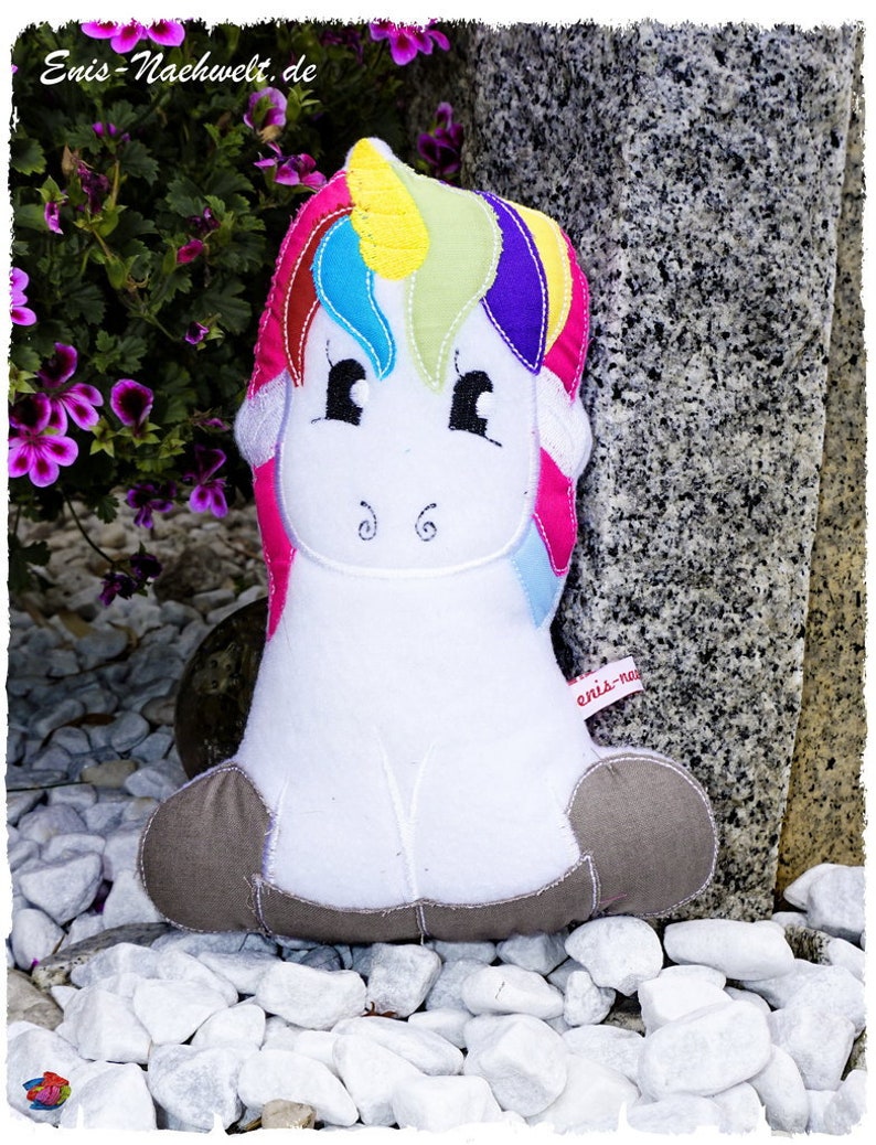 May include: A plush unicorn toy with a white body, gray hooves, and a colorful mane and tail. The mane features sections of yellow, blue, green, and purple. The toy has black eyes and embroidered details. The text "enis-nachwelt.de" is visible.