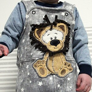 May include: A gray denim overall with white stars and a brown lion applique. The lion has a brown mane and a white face with black eyes and a black nose.