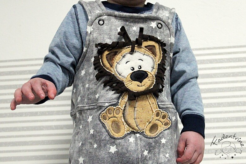 May include: A gray denim overall with white stars and a brown lion applique. The lion has a brown mane and a white face with black eyes and a black nose.