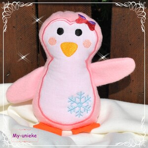 May include: A pink plush penguin with a light blue snowflake design on its chest. The penguin has black eyes, orange feet, and a yellow beak. A red and purple bow adorns its head. The text "My-unieke" is visible at the bottom.