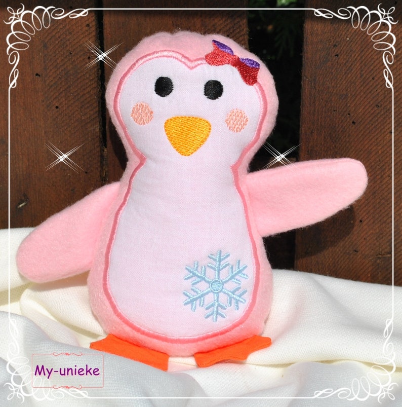 May include: A pink plush penguin with a light blue snowflake design on its chest. The penguin has black eyes, orange feet, and a yellow beak. A red and purple bow adorns its head. The text "My-unieke" is visible at the bottom.