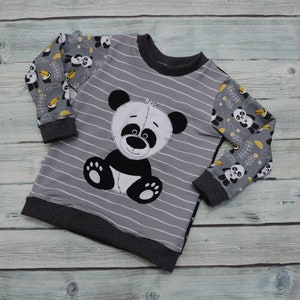 May include: A gray and black striped long-sleeved shirt with a panda bear design on the front. The sleeves are patterned with small panda bears and yellow leaves.