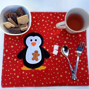 May include: A red placemat with gold stars features a penguin applique holding a gingerbread man. A white bowl of cookies, a mug of tea, sugar cubes, and silverware complete the festive table setting.
