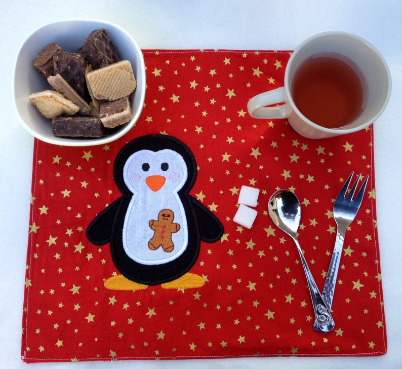 May include: A red placemat with gold stars features a penguin applique holding a gingerbread man. A white bowl of cookies, a mug of tea, sugar cubes, and silverware complete the festive table setting.