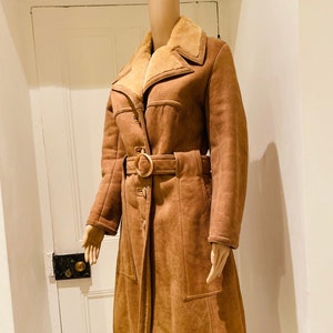 May include: A brown suede coat with a shearling collar and a belt. The coat has a long, fitted silhouette and a button closure.