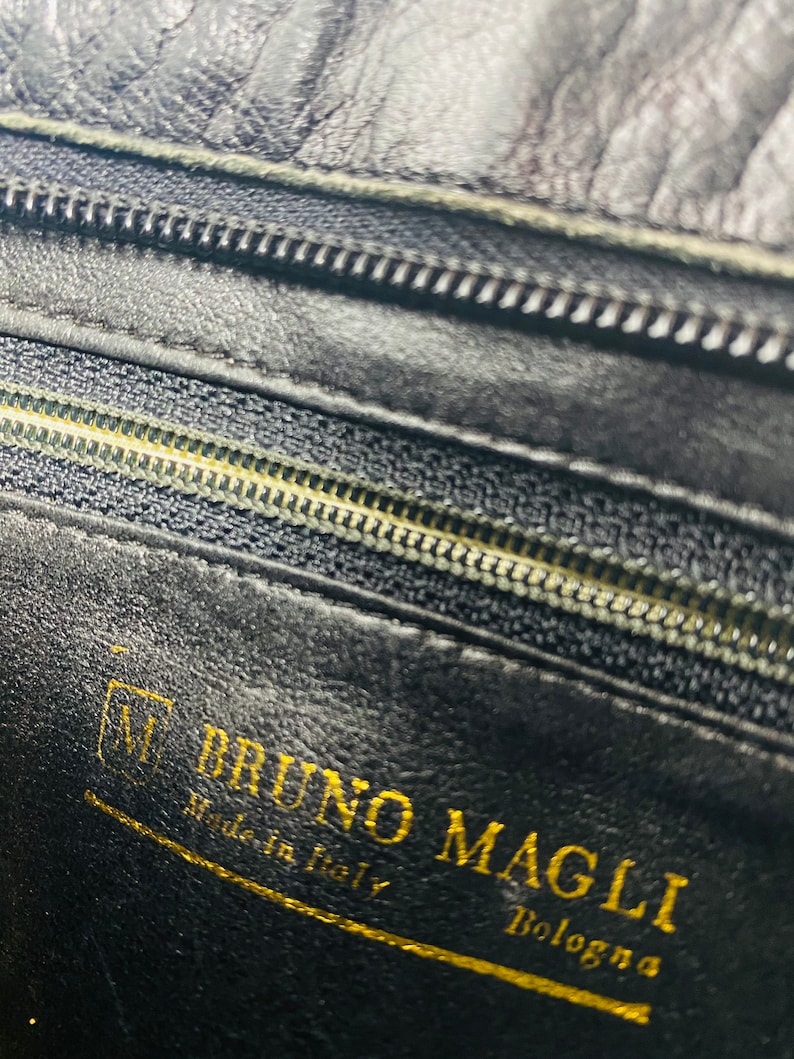 May include: Close-up of a black leather interior with a gold zipper and a gold label that reads "BRUNO MAGLI Made in Italy Bologna".