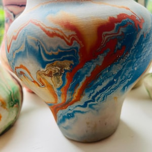 May include: A ceramic vase featuring a marbled pattern with swirling colours of bright blue, vibrant orange, and muted gold against a white base. The vessel has a rounded, bulbous body that tapers towards the top opening, showcasing a smooth finish.