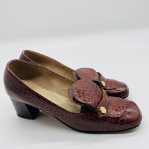 May include: A pair of brown leather heeled shoes with a crocodile pattern. The shoes feature a gold buckle detail and a low block heel.