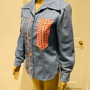 May include: Pale blue denim shirt with a collared neckline and long sleeves. It has a red and white gingham pocket and zip detail, plus blue and white checked accents. The shirt has white buttons and decorative stitching.