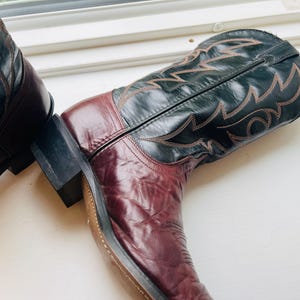 May include: A pair of two-toned cowboy boots featuring a deep burgundy leather foot and a black leather shaft. The black shaft is decorated with intricate, light-coloured stitching in a flame-like pattern. Each boot has a stacked heel.