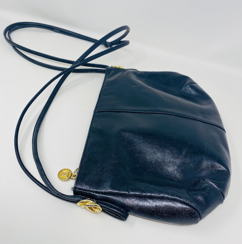 May include: A black leather shoulder bag with a gold clasp and a gold logo charm. The bag has a long strap and a rounded shape.