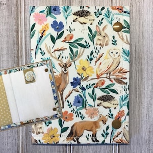 May include: A fabric journal cover and matching insert featuring a woodland animal and floral pattern. The cover has a white background with illustrations of a deer, fox, owl, rabbit, and hedgehog, along with colorful flowers and foliage.
